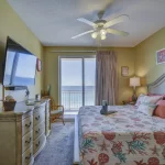 Guest bedroom painted yellow with light wood furniture including a bed, nightstand, and dresser. A balcony overlooks the ocean