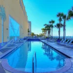Hotel pool surrounded by beach chairs and palm trees near the ocean. The wall of the hotel has palm trees painted on it