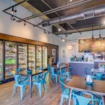 Cafe store including seating areas with blue chairs, drink refrigerators, a counter, and blackboard menu