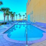 Hotel pool surrounded by beach chairs and palm trees near the ocean. The wall of the hotel has palm trees painted on it