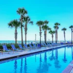 Hotel pool surrounded by beach chairs and palm trees near the ocean